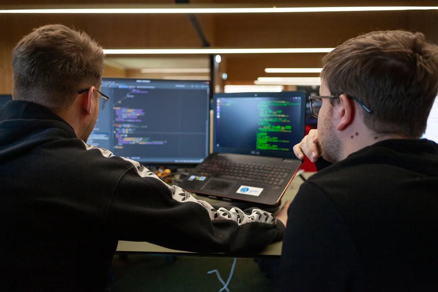Two developers sitting together at a desk, working through code shown on a laptop and a second monitor