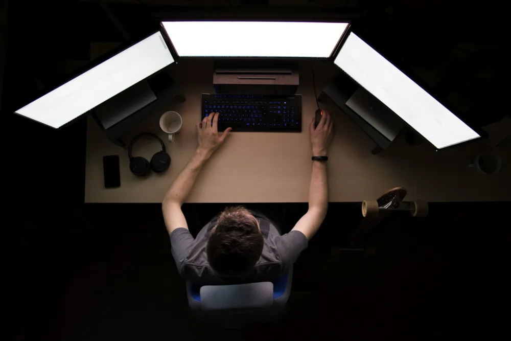 Overhead view of a developer working at a desk under three lit monitors in a dark room